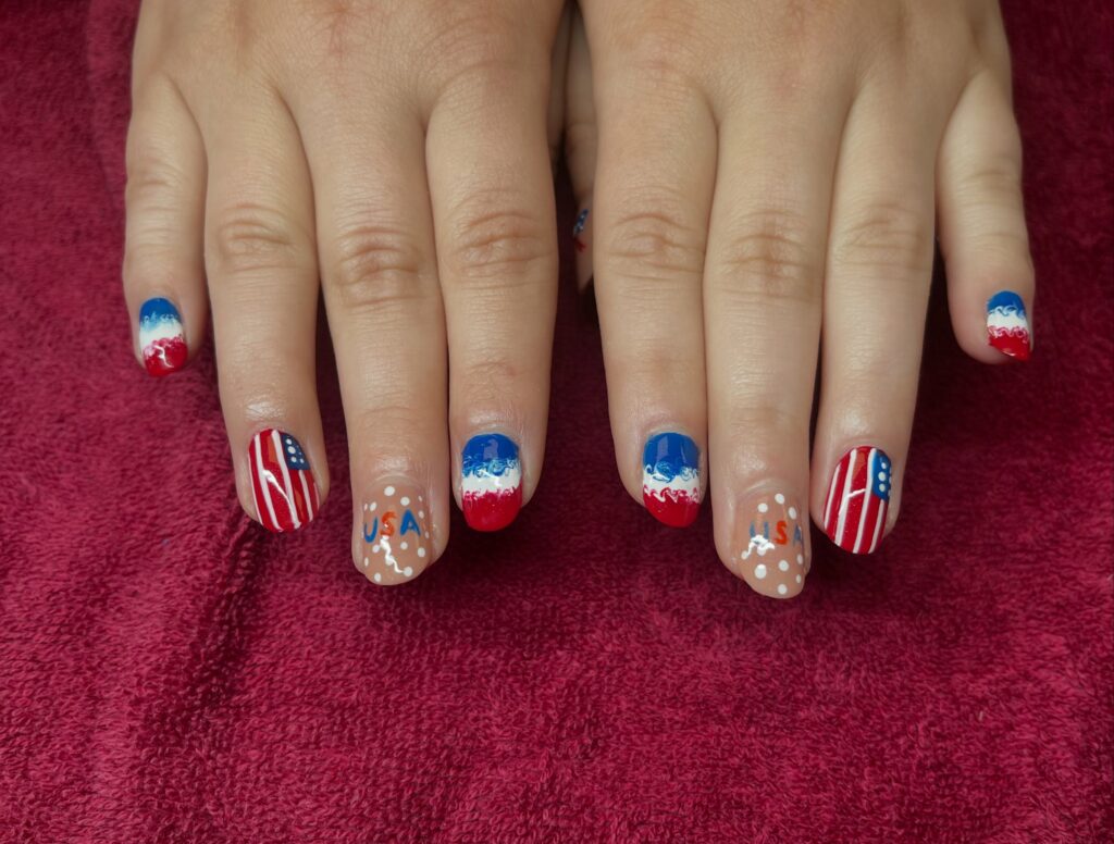 Patriotic nail art with red, white, and blue designs featuring American flags, USA lettering, stars, and decorative patterns.